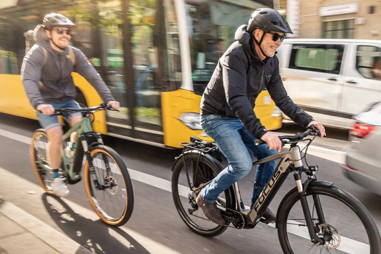 Zwei E-Biker fahren auf E-Bikes von Velocultour auf einem Fahrradstreifen in der Innenstadt. Im Hintergrund ein Bus und mehrere Autos.